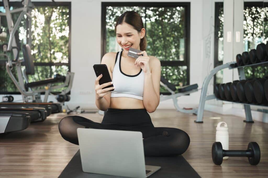 Contact Diboll Fitness | Happy Woman Using Mobile Phone And Credit Card For Online Shopping In Gym.